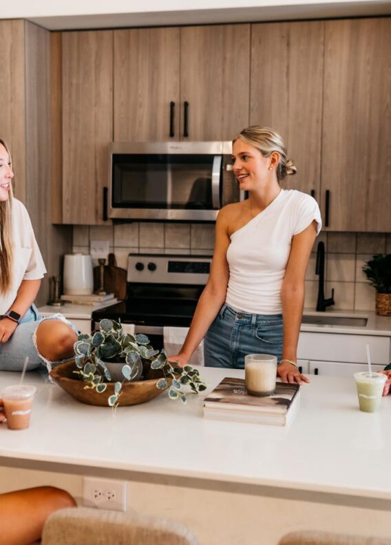 Four women converse and laugh around a modern kitchen island, each with a drink in hand. The kitchen features wood cabinets, stainless steel appliances, and a decorative plant centerpiece on the white countertop.
